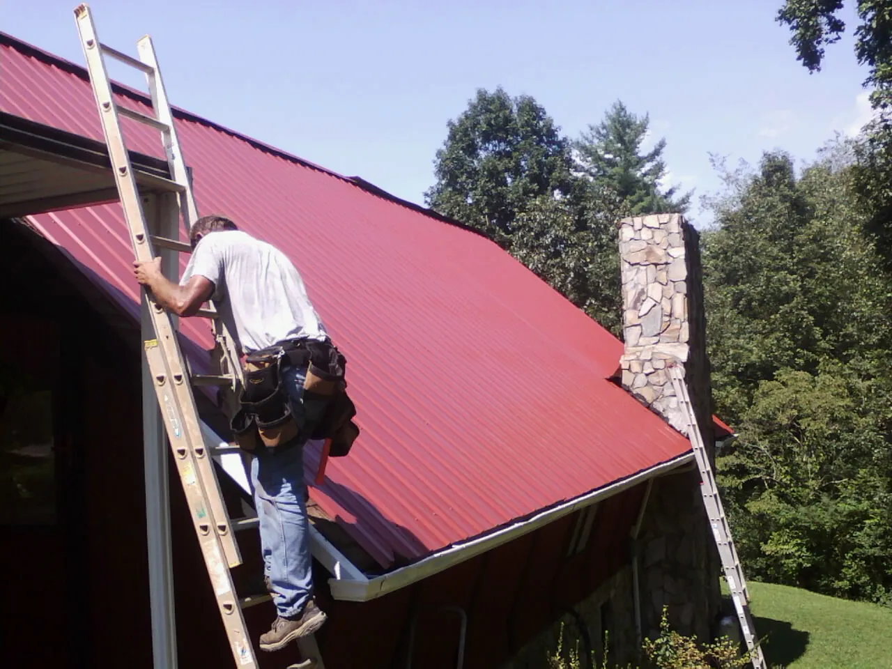 Roofer on a ladder beside a red metal roof and stone chimney — Soffit & Fascia Repair by Roofing Brothers in Emory